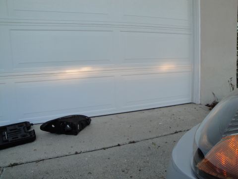 2003 Ford Taurus - A detached headlight assembly rests on the ground near a garage door.