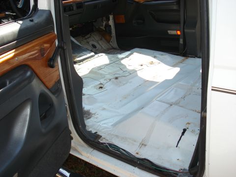 1992 Ford C-350 (Centurion Conversions) - View through the open driver's door of a white extended-cab Ford truck showing the bare white floor pan with cracked seam sealer and rust s…