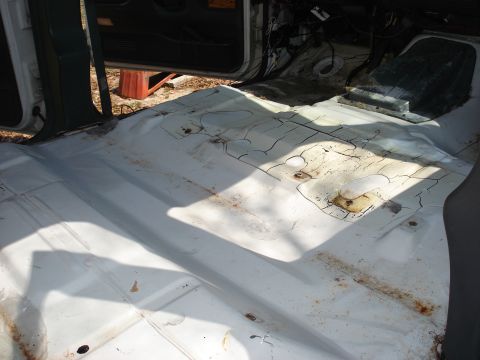 1992 Ford C-350 (Centurion Conversions) - Bare floor pan of a stripped truck cab shot from the open passenger door, painted white with surface rust streaks and cracked, peeling seam…