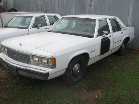 1991 Ford Crown Victoria LTD - Two white sedans are parked outside next to a corrugated metal fence. One has a black patch on the door.