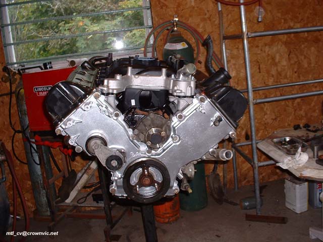 Aluminum V engine on a stand in a workshop with welder and wood-paneled wall behind