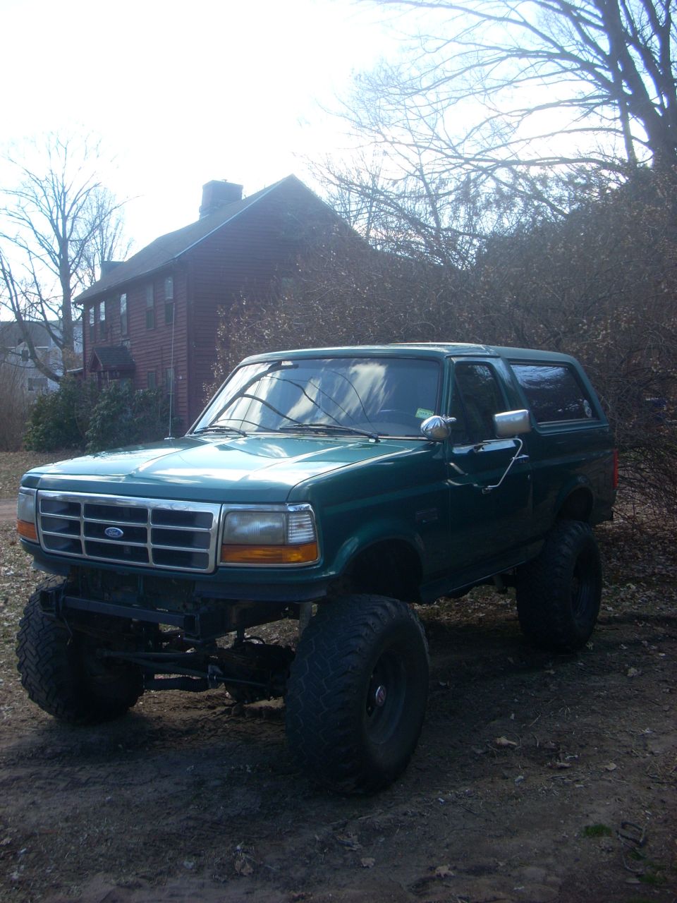 Lifted teal-green Ford Bronco with front bumper removed, exposed axle, parked on dirt