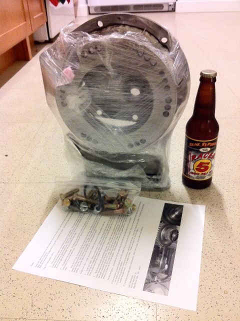 1989 Ford Bronco - A transfer case adapter plate, hardware, and a bottle of beer are laid out on a speckled floor.