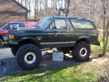 1989 Ford Bronco - A lifted dark green Ford Bronco with oversized tires sits parked in a grassy area next to a red vehicle.