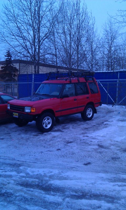 1990 Chevrolet Suburban - A red SUV with a roof rack sits in a snowy lot next to another vehicle.