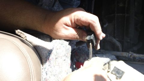 1994 Ford Bronco - Hands hold a small black electrical connector and probe it with a screwdriver-like tool over a peeled-back floor area, exposing gray jute c…