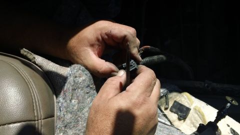 1994 Ford Bronco - Two hands use a small pick to release a terminal from a connector on a conduit-wrapped wiring harness inside a vehicle with the carpet pull…
