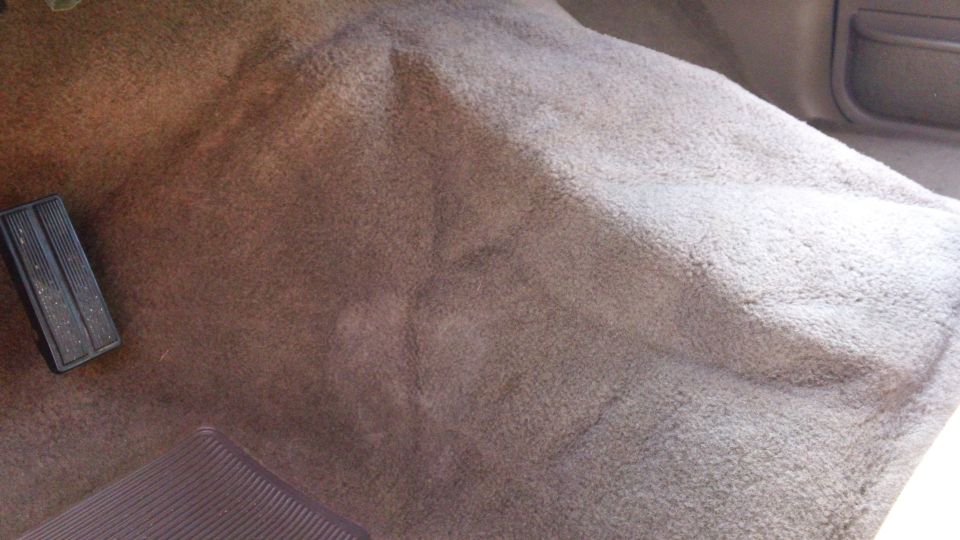 Car interior floor with brown carpet and black pedals. Water stain visible
