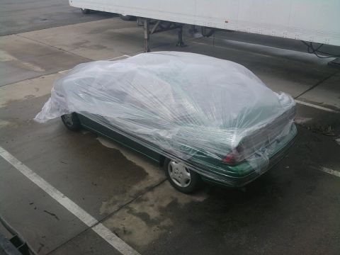 1989 Ford Bronco - Car covered in plastic wrap, parked outdoors near a trailer.