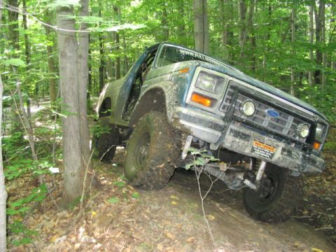 1986 Ford Bronco - A green Ford Bronco navigates a muddy, wooded trail at an angle.