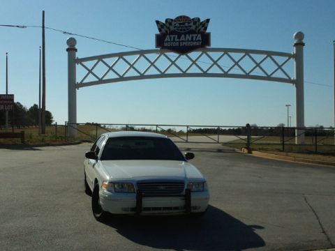 1998 Ford Crown Victoria - A white 1998 Ford Crown Victoria, equipped with a push bar, is parked in front of the Atlanta Motor Speedway entrance gate.