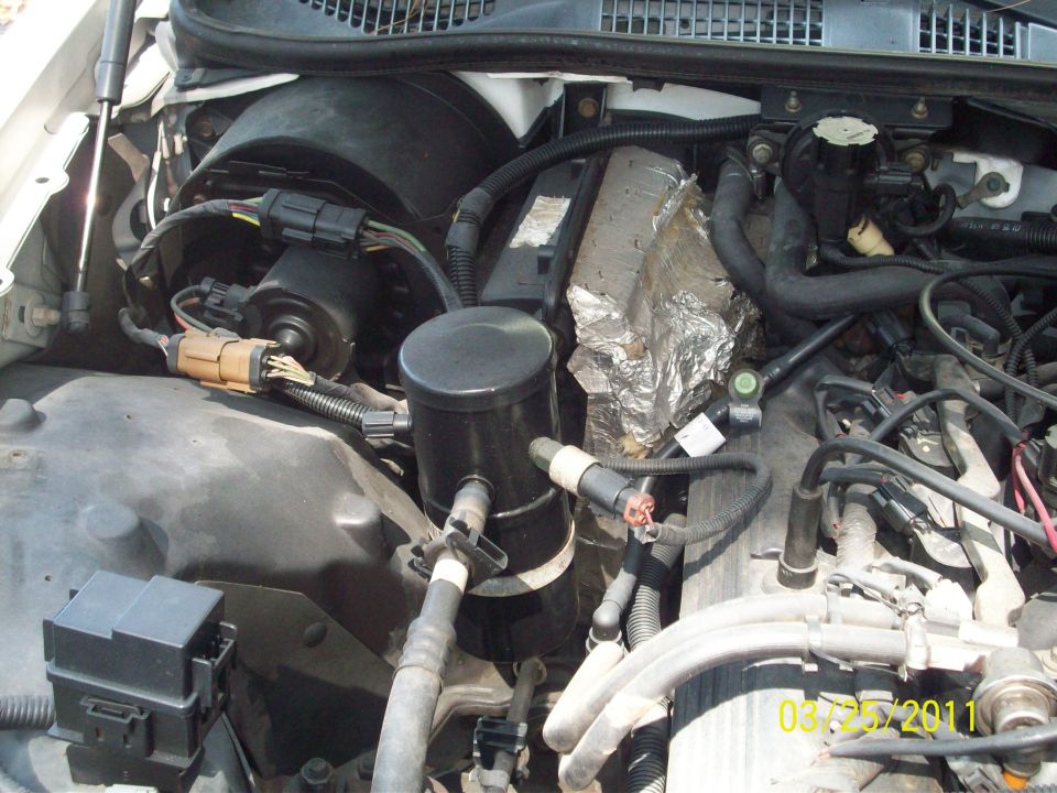 Engine bay of white car with black canister, wiring, hoses, and foil-wrapped component; dated 03/25/2011