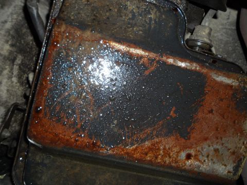 1989 Ford Bronco - Close-up of a heavily corroded and oil-stained automotive part, likely an oil pan, with a watermark.