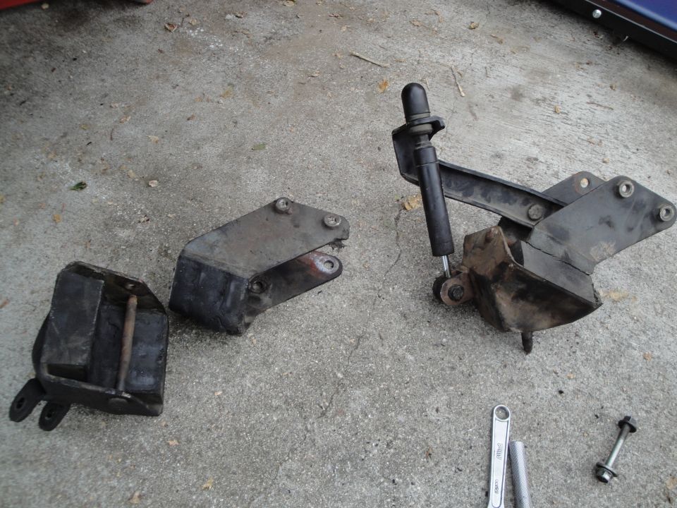 Three removed steel hood hinges on concrete, one with gas strut, plus wrenches and bolts