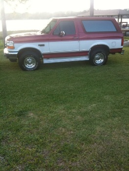 1995 Ford Bronco - A two-tone red and white Ford Bronco is shown from the side, parked on a grassy area next to a body of water.