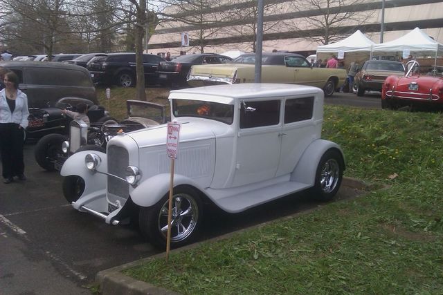 White sedan hot rod with chrome wheels and a tall grille, parked on grass