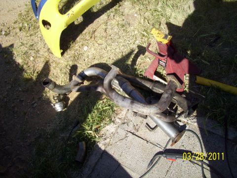 1995 Ford Probe - Exhaust system parts are laid out on the ground with a yellow object and a red jack stand nearby.