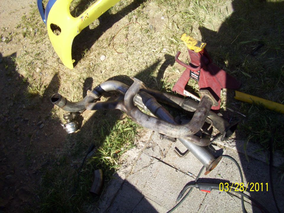 Exhaust manifold and pipes on the ground next to a yellow plastic object and a red jack stand
