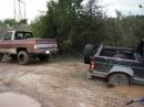 1989 Ford Bronco - Two off-road vehicles are stuck in the mud on a dirt path, surrounded by trees and vegetation.