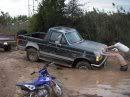 1989 Ford Bronco - A person attempts to push a dark pickup truck out of deep mud, while a motorcycle sits in the foreground.