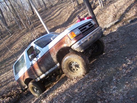 1989 Ford Bronco - A muddy Ford Bronco navigates a challenging off-road trail in a wooded area.