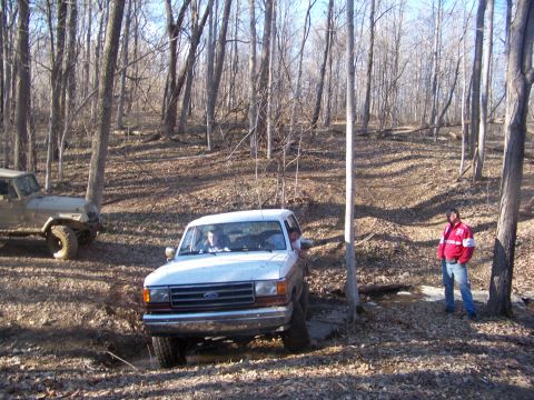 1989 Ford Bronco - A white Ford truck navigates a muddy trail in a wooded area with another vehicle visible.