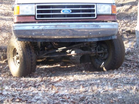 1989 Ford Bronco - This lifted Ford Bronco is shown from the front, with its grille and bumper prominent. The vehicle is on a dirt and leaf-covered surface.
