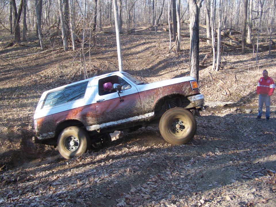 1989 Ford Bronco climbing muddy hill in wooded area. Person stands watching