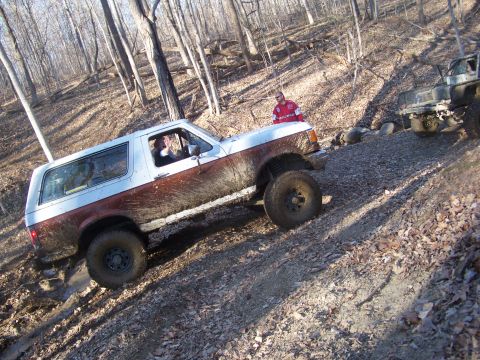 1989 Ford Bronco - A white and brown Ford Bronco navigates a muddy, wooded trail with another off-road vehicle in the background.