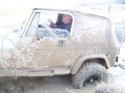 1989 Ford Bronco - A mud-caked Jeep Wrangler navigates through a watery, muddy terrain with a driver behind the wheel.