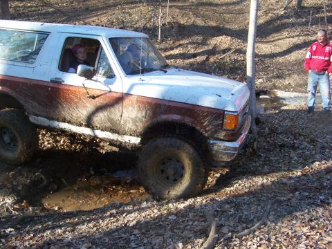 1989 Ford Bronco - A white and brown Ford Bronco navigates a muddy trail with a person in the driver's seat and another standing nearby.