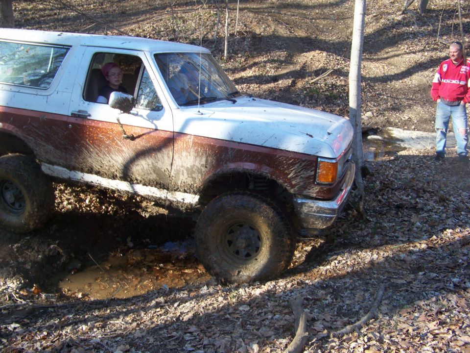 Mud-splattered white and brown Ford Bronco driving through a muddy wooded area