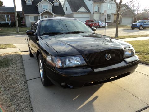 2004 Mercury Grand Marquis - Black sedan with a blacked-out grille and Mercury logo, parked on a driveway with houses in the background.