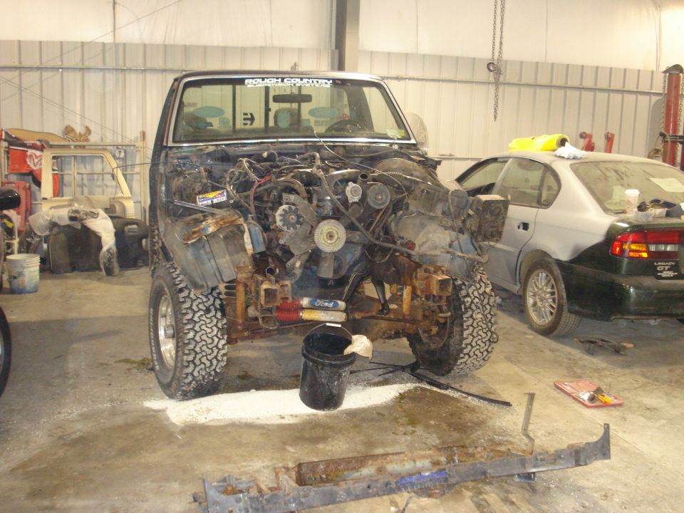 Truck with front end removed, showing engine and chassis. Off-road tires. "ROUGH COUNTRY" sticker on windshield