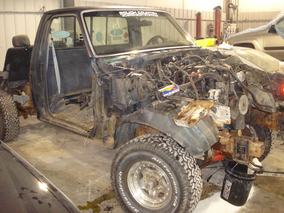 Disassembled truck in a garage, engine bay empty, doors open. Rough Country sticker on windshield