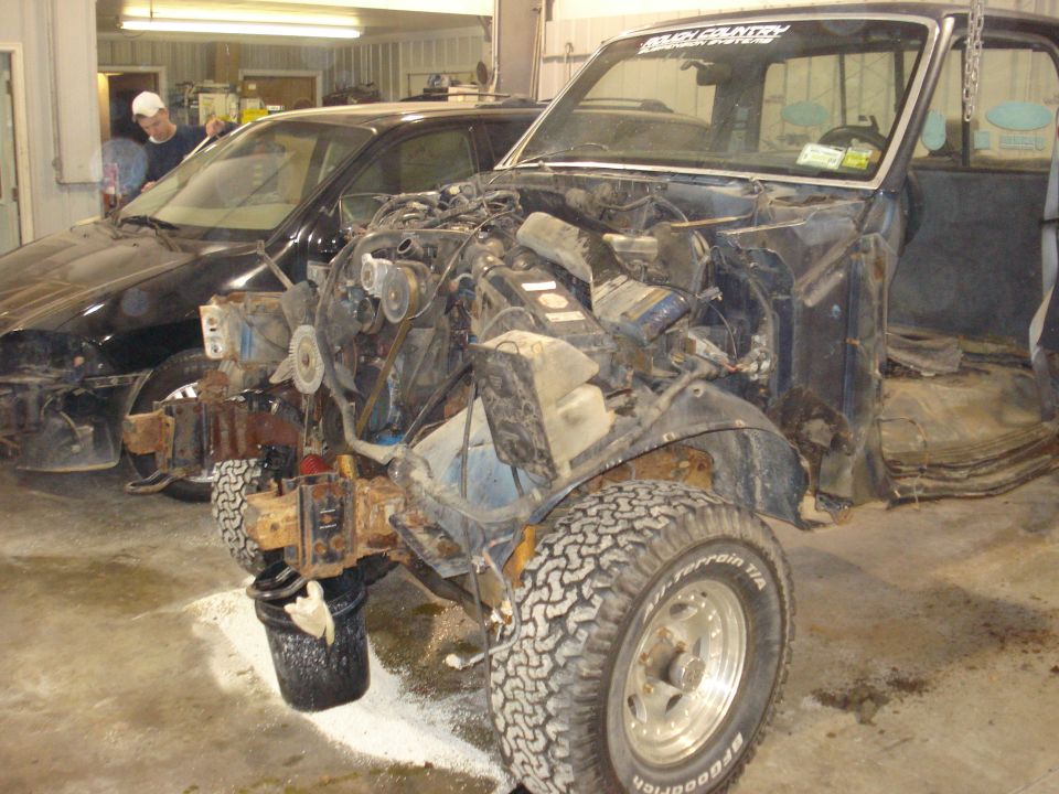 Truck stripped down, engine out, parked next to a black car in a garage