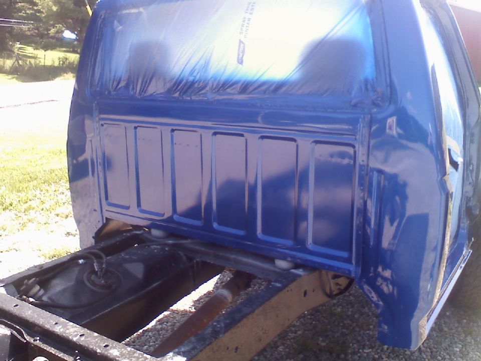 Rear view of a blue pickup truck bed with plastic sheeting covering the rear window area. The frame and fuel tank are visible