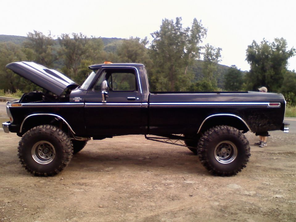 Side view of a black lifted pickup truck with the hood open. It has large off-road tires and chrome trim