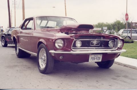 1981 Ford Bronco - A modified 1967 Ford Mustang with a prominent hood scoop sits parked on pavement, with another car visible behind it.