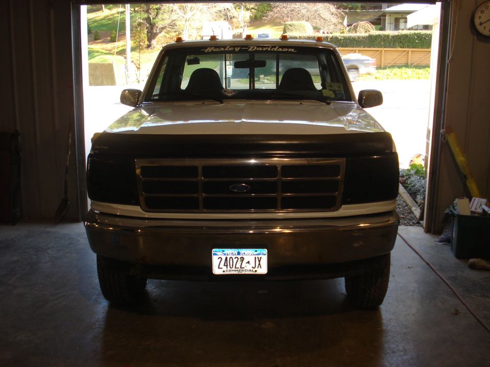 Front view of a white Ford F-150 truck with a black grille guard and Harley-Davidson banner on the windshield. License plate reads 24022-JX