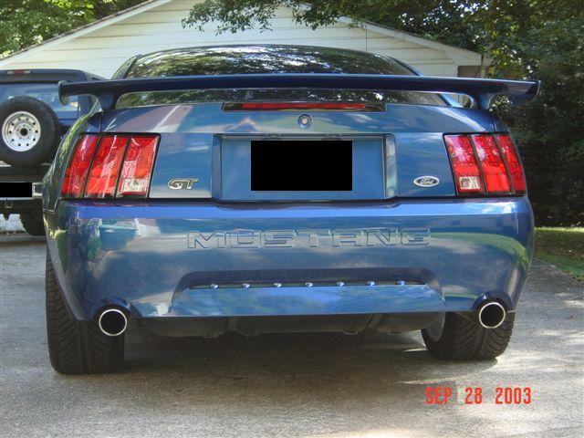 Rear view of blue Mustang GT with spoiler and dual exhaust. A truck is parked to the left