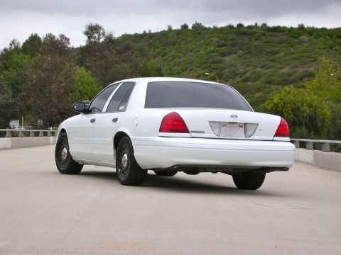 2004 Ford Crown Victoria - White Ford Crown Victoria sedan with steel wheels, photographed from the rear quarter on a road bordered by green hillsides.