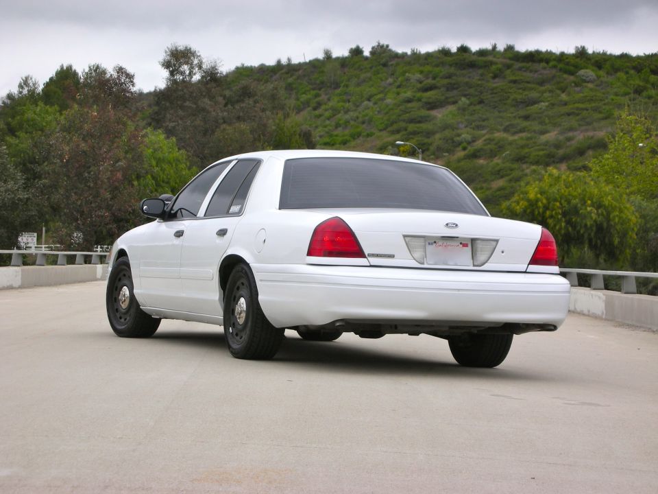 Rear three-quarter view of white Ford Crown Victoria sedan on a road by green hills