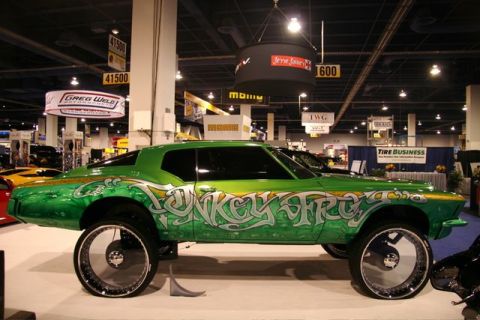 1991 Ford LTD Crown Victoria - Green candy-painted 1970s Buick Riviera boat-tail coupe on a raised display stand at an indoor trade show, sitting on oversized chrome mult…