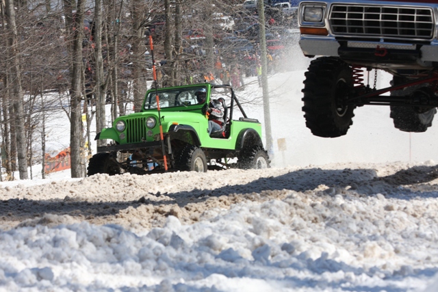 Green Jeep and a lifted Ford truck mid-air over snow. The Ford truck's grille is visible