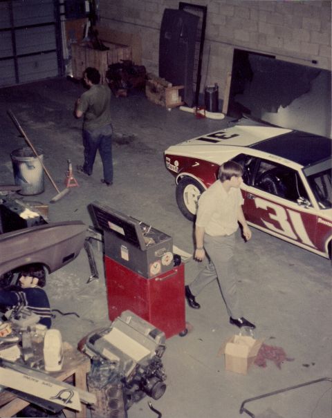 1973 Ford Mustang - A race car with prominent numbering is seen in a garage setting, with a person nearby and other automotive parts scattered around.