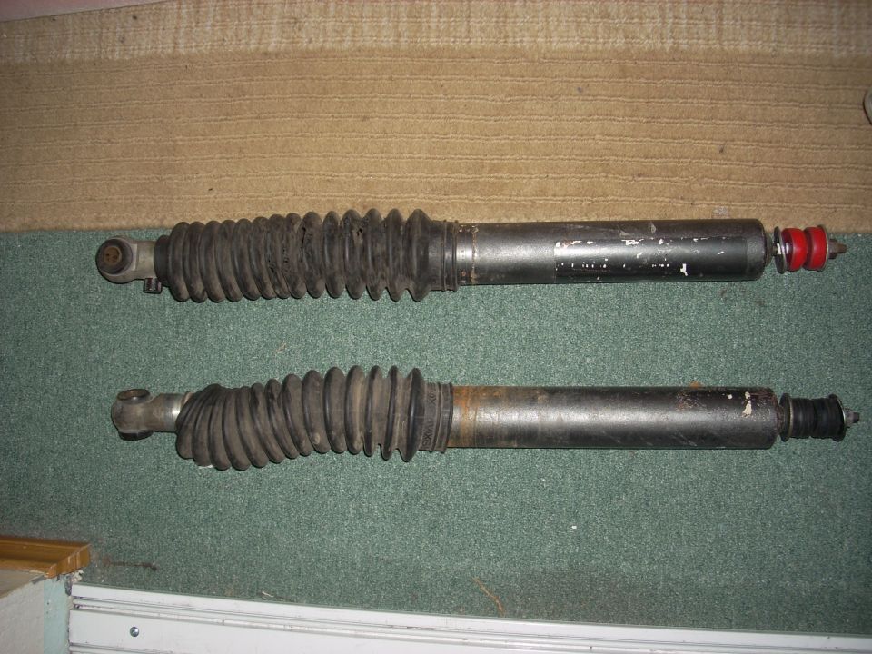 Two black shock absorbers with rubber bellows, one with red end caps, on a carpeted surface