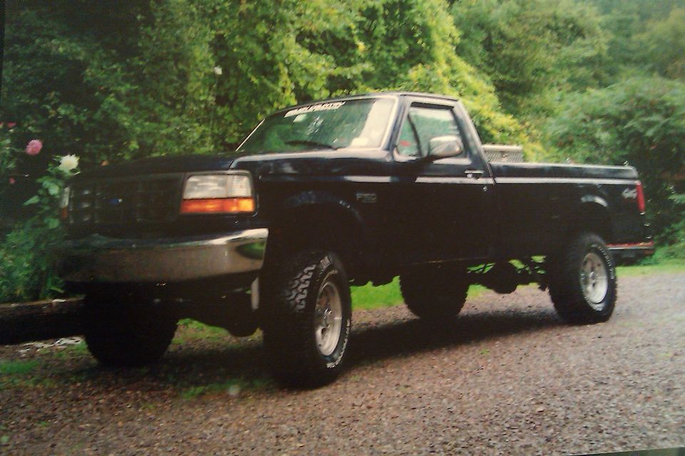 Black Ford F-150 pickup truck, lifted, with chrome bumper and aftermarket wheels, parked on a gravel surface