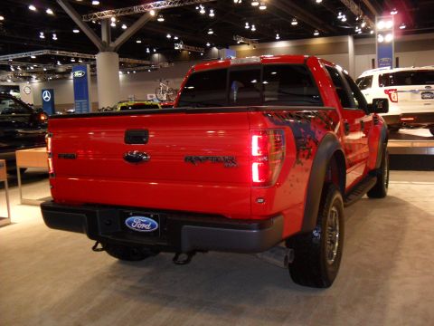 2011 Ford F-150 - A red Ford F-150 Raptor pickup truck is displayed indoors, with another vehicle visible in the background.