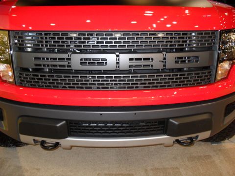 2011 Ford F-150 - Close-up of the front grille and bumper of a red Ford F-150 Raptor, showing the 'FORD' emblem.